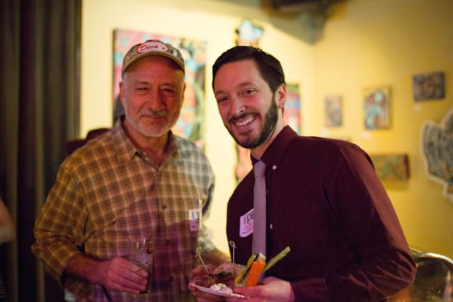 Two men smiling together at a social event, one holding a drink and the other a plate of food, with colorful artwork in the background, capturing the communal spirit of the Love Bomb 2017 event supporting My Voice Music.